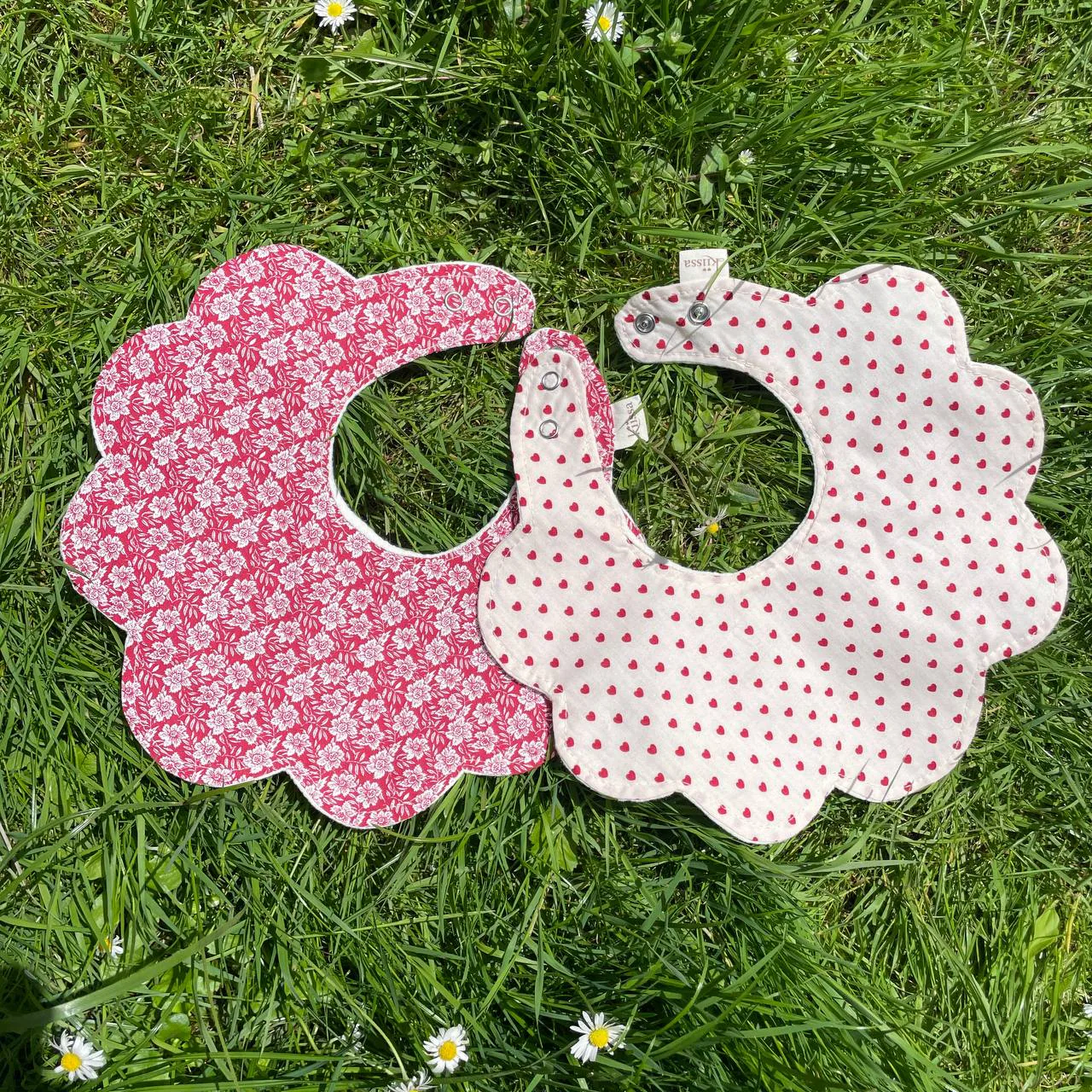 A red floral bib and a hearts bib side by side on the grass
