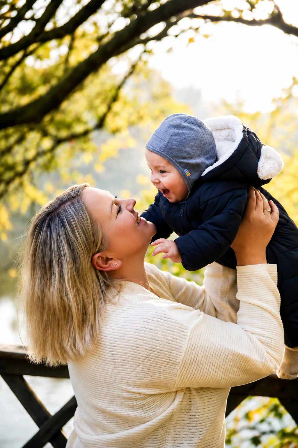 A mother lifting her smiling baby into the air under autumn trees